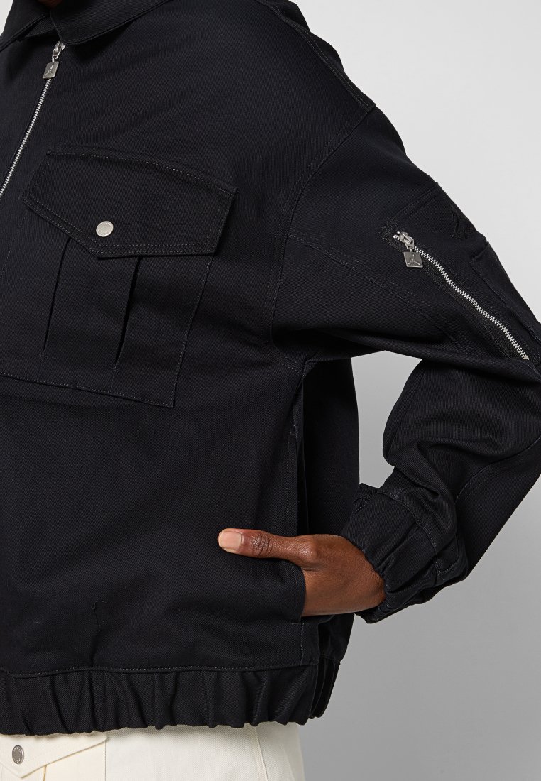 Giacca in denim nera con zip frontale, due tasche sul petto e una tasca con zip aggiuntiva sulla manica. Presenta un orlo e polsini a coste.