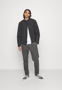 Gepolsterte schwarze Jacke mit hohem Kragen und Reißverschluss, kombiniert mit grauen Hosen und blauen Sneakers mit weißen Streifen. Darunter ein schlichtes weißes T-Shirt.
