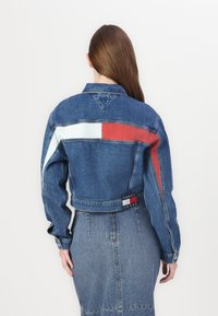 Mėlynai nukirptas džinsinis švarkas su raudonais ir šviesiai mėlynais akcentais. Yra etiketė su užrašu "TOMMY JEANS" ir klasikinis apykaklė.