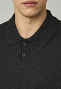 Polo shirt noir avec un tissu texturé, trois boutons noirs au col et un design de col classique.