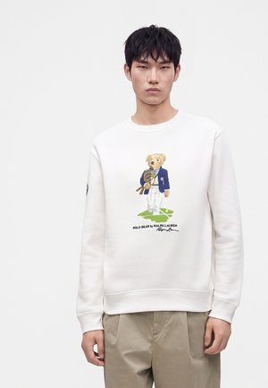 Jeune homme portant un sweat-shirt blanc avec un graphisme d'ours en blazer bleu tenant des raquettes de tennis et un pantalon beige.