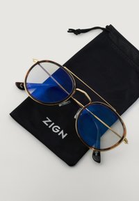 Ronde zonnebrillen met blauwgetinte lenzen, gouden metalen frame en bruin schildpadaccenten, liggend op een zwarte zak met het "ZIGN" logo.