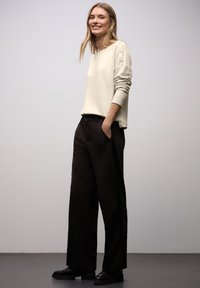 Kremowy sweter z ozdobnymi guzikami na ramieniu, w połączeniu z luźnymi ciemnymi spodniami i czarnym obuwiem; ogólnie gładka faktura.
