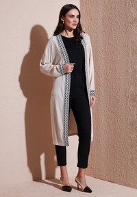 Long cream cardigan with black embroidered trim, worn over a black jumpsuit and paired with black and clear heeled shoes. Textured fabric.