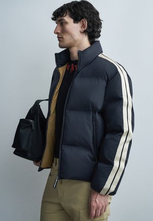 Mladý muž s čiernymi kučeravými vlasmi, oblečený v tmavomodrej prešívanej bunde s bielymi pásmi, béžových nohaviciach a s čiernym batohom.