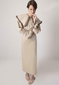 Γυναίκα που φοράει μακρύ μπεζ φόρεμα με μεγάλο βολάν στο γιακά και στα μανίκια, στέκεται με το χέρι κοντά στο στόμα σε απλό φόντο.