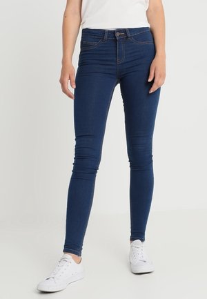 Slim fit dżinsy w ciemnoniebieskim kolorze, z wysokim stanem, dyskretnymi przeszyciami i dopasowanym krojem. W połączeniu z białymi sneakersami.