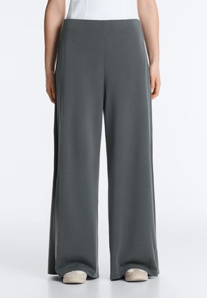 Personne portant un pantalon large gris ample et des chaussures blanches, debout contre un fond uni, les mains détendues le long du corps.