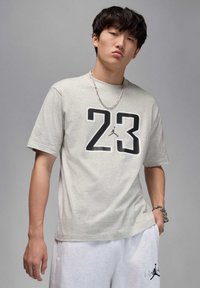 Jasnoszara koszulka z krótkim rękawem z czarnym numerem "23" i logo Jumpman na przodzie. Miękka tkanina, luźny krój, w zestawie z białymi szortami.