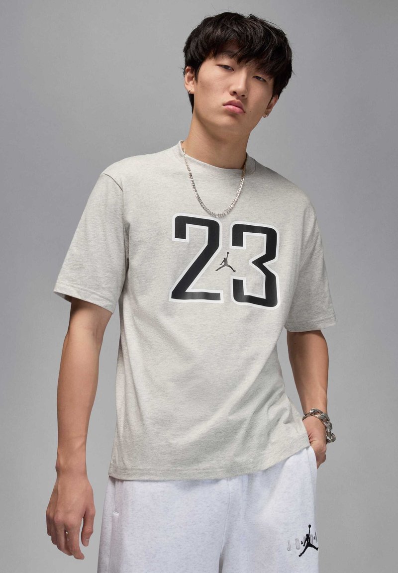 Jasnoszara koszulka z krótkim rękawem z czarnym numerem "23" i logo Jumpman na przodzie. Miękka tkanina, luźny krój, w zestawie z białymi szortami.