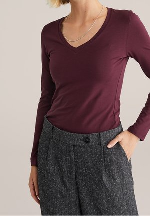 Vrouw draagt een aansluitend, donkerpaars, lang mouw topje met V-hals, ingestopt in een grijze, hoog getailleerde tweedbroek met knopen aan de voorkant en hand in de zak.