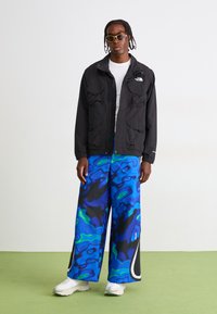 Mand med sort North Face-jakke, hvid skjorte, blå brede bukser med abstrakt mønster, hvide sneakers og gule solbriller står på grønt gulv.