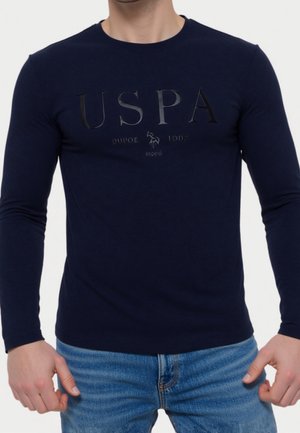 Homme portant une chemise à manches longues bleu marine avec logo USPA en relief et graphique, associée à un jean bleu, debout avec les mains près des hanches.