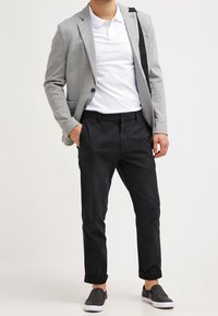 Blazer gris claro sobre una polo blanca, combinado con pantalones negros arremangados y zapatos oscuros de tipo slip-on, creando un look casual pero estructurado.