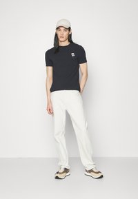 T-shirt blu navy con piccolo logo bianco, abbinata a pantaloni bianchi e sneakers beige. Il modello indossa un cappellino beige chiaro. Outfit semplice e casual.