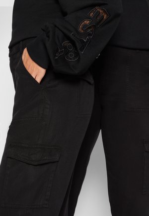 Person wearing black cargo pants with flap pockets and black sweatshirt with ESA 87 embroidery on sleeve, hand in pocket.