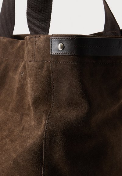 Sac cabas en daim brun foncé avec garniture en cuir noir et rivet argenté, avec des bretelles d'épaule en tissu tissé noir.