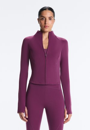 Woman with dark hair tied back wears a fitted purple zip-up long-sleeve jacket and matching leggings, standing against a white background.