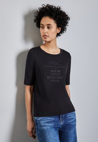 T-shirt noir à manches courtes, avec des lettres texturées blanches portant le mot "PRINTEMPS" et des phrases détaillées, associé à un jean bleu.