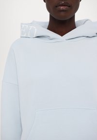 Personne portant un sweat à capuche bleu clair avec le texte blanc "ED" sur la capuche, montrant la partie supérieure de la poitrine et le cou sur un fond uni.