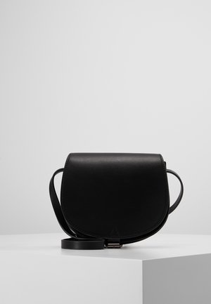 Sacoche en cuir noir avec fermeture à rabat et bandoulière réglable, posée sur un socle blanc devant un fond gris.