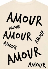 T-shirt beige avec du texte noir portant le mot "AMOUR" dans différentes tailles et orientations, créant un motif dynamique sur le tissu.