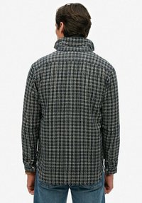 Man in een blauw-wit geruite lange mouw shirt met hoge kraag, gezien van achteren, gecombineerd met blauwe jeans.