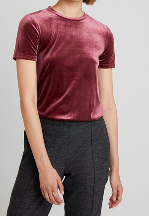 Persoon draagt een kortärmlige, stofroze fluwelen top en een hoge taille gestructureerde donkergrijze broek, staat met ontspannen armen.