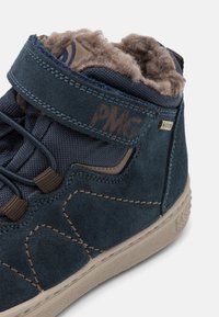 Bottines montantes en suede bleu marine avec une doublure douce, une semelle en caoutchouc et des accents contrastants en tan. Présente une sangle avec logo et un tissu texturé.