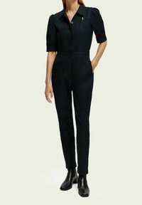 Vrouw in een zwarte lange jumpsuit met zakken, knoopsluiting aan de voorkant en zwarte haklaarzen, staand tegen een effen achtergrond.