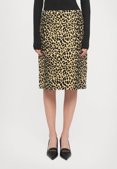 Woman wearing a knee-length leopard print skirt, black long-sleeve top, and black pointed-toe shoes with gold detailing, standing on white background.