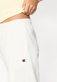 Λευκός παντελόνι jogging με ελαστική μέση και πλαϊνές τσέπες· διαθέτει ένα μικρό, κεντητό λογότυπο σε μπλε, κόκκινο και λευκό.