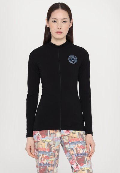 Camiseta negra de manga larga con cremallera y cuello alto, que presenta un logo circular azul en el lado izquierdo del pecho, combinada con pantalones de estampado.