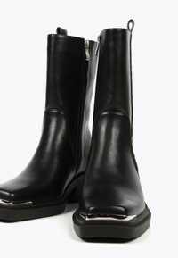 Black leather ankle boots with a square toe, side zippers, and a shiny metallic accent on the toe. Smooth texture and sturdy sole.