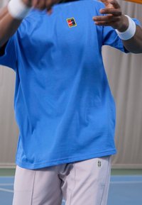 Person wearing blue Nike tennis shirt and white shorts with wristbands, preparing to hit a tennis ball on an indoor court.