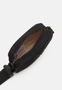 Sort nylon pouch med lynlåslukning, beige indvendig foring og en robust rem med spænde. Subtil tekstur og minimalistisch design.