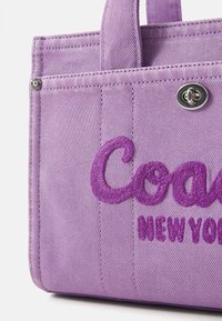 Purple fabric tote bag featuring embroidered "Coach" and "New York". Textured surface with contrasting stitching and a circular silver snap hardware.