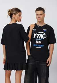Sorte T-shirts med grafiske designs, der præsenterer fed hvid og blå tekst, båret af to modeller; den ene har også en sort plisseret nederdel på.