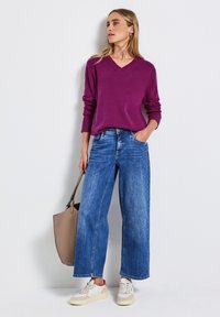 Un pull violet en V, un jean bleu à jambes larges et des baskets beiges. Tient un sac fourre-tout marron clair, avec un fond uni.