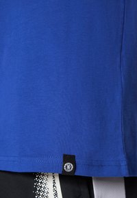 Blå bomulds-t-shirt med en glat tekstur, der har et lille Chelsea FC-logo ved kanten og en simpel rund halsudskæring.