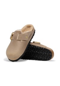 Sabots beiges avec doublure en fausse fourrure, sangle ajustable et détails en métal doré. Semelles en caoutchouc noir texturé pour une meilleure adhérence. Nom de la marque en relief.