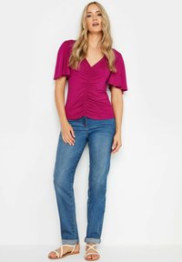 Top fucsia con mangas flotantes y fruncido en el frente; combinado con jeans de mezclilla de lavado medio; la modelo lleva sandalias blancas de tiras.