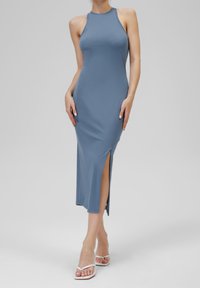 Bezramienna, jasnoniebieska midi sukienka z elastycznego materiału, z bocznym rozcięciem i dopasowanym krojem. Noszona z białymi sandałami.