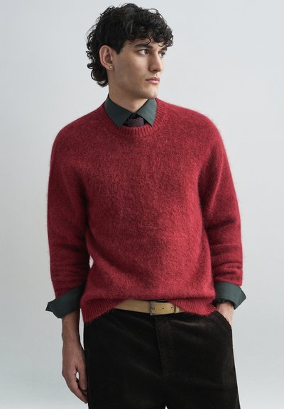 Pull en maille rouge flou à col rond, porté sur une chemise verte foncée à col. Pantalons en velours côtelé foncé et ceinture beigne visibles.