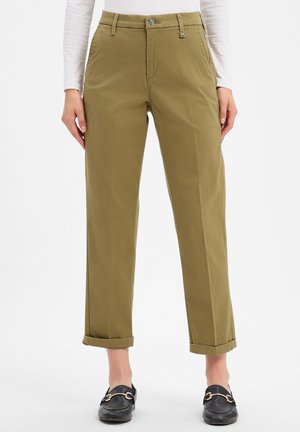 Personne portant un pantalon droit vert olive avec des revers, associé à des chaussures noires sans lacets et un haut blanc à manches longues.