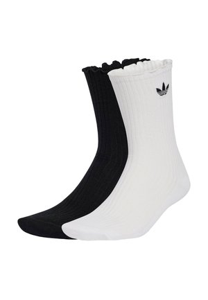 Jedna czarna i jedna biała prążkowana skarpetka za kostkę z falbankowymi mankietami, na białej skarpetce znajduje się małe czarne logo Adidas trefoil blisko góry.