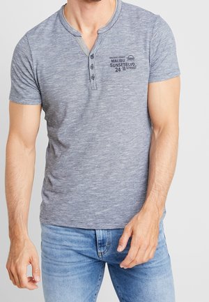 Szary, prążkowany T-shirt typu henley z krótkim rękawem, dekoltem zapinanym na guziki i nadrukiem na lewej piersi. Tkanina wydaje się miękka i gładka.