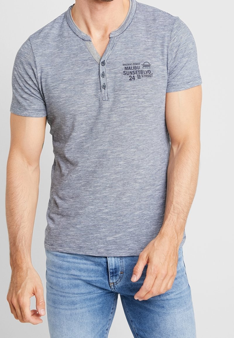 Chemise henley à manches courtes rayée grise avec un col à boutons et un détail de texte imprimé sur le côté gauche de la poitrine. Le tissu semble doux et lisse.