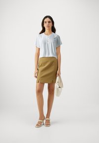 Blusa azzurra chiara a maniche corte con pinces e dettaglio dei bottoni, abbinata a una gonna aderente gialla senape con spacco laterale. Accessoriata con una borsa bianca.
