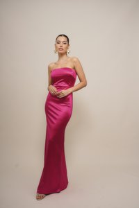 Woman with pulled-back hair wearing large gold earrings and a strapless bright pink satin gown, standing against a plain beige background.